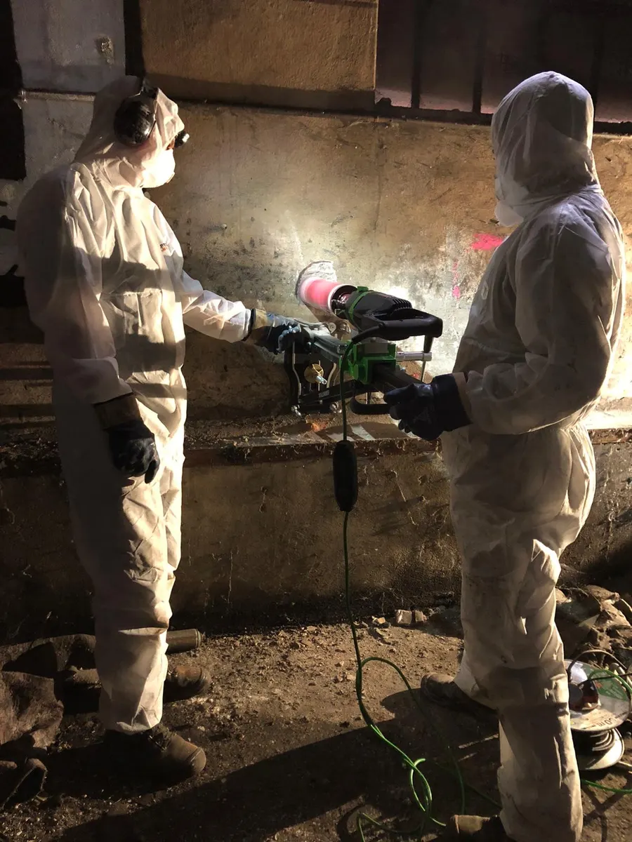 Zwei Mitarbeiter von TERRA V in Schutzanzügen führen eine Kernbohrung in der Bausubstanz eines Kellers aus